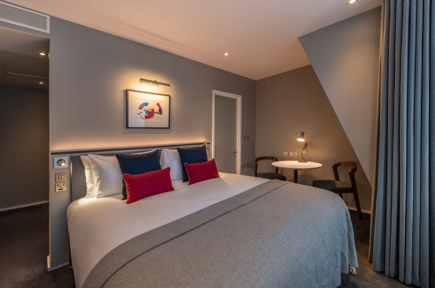 A modern hotel room with a large bed, gray walls, blue and red accent pillows, a small round table with two chairs, framed artwork above the bed, and floor-to-ceiling curtains on the right.