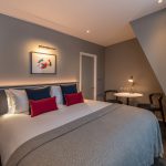 A modern hotel room with a large bed, gray walls, blue and red accent pillows, a small round table with two chairs, framed artwork above the bed, and floor-to-ceiling curtains on the right.