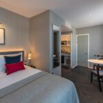 A modern hotel room with a neatly made double bed, colorful pillows, gray bedding, a small dining area with chairs, kitchenette, wall art, large window with curtains, and neutral-colored walls and carpet.