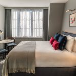 Modern hotel room with a large bed, white linens, and colorful pillows. A small round table with two chairs sits by a window with sheer curtains. Minimalist decor includes a framed abstract artwork above the bed.