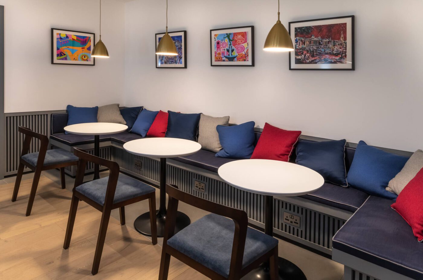 A cozy cafe corner at The Resident Covent Garden features a long cushioned bench with blue, grey, and red pillows lining the wall. White round tables and dark wooden chairs are neatly arranged. Three colourful framed artworks hang on the wall above the seating area, and pendant lights hang overhead.