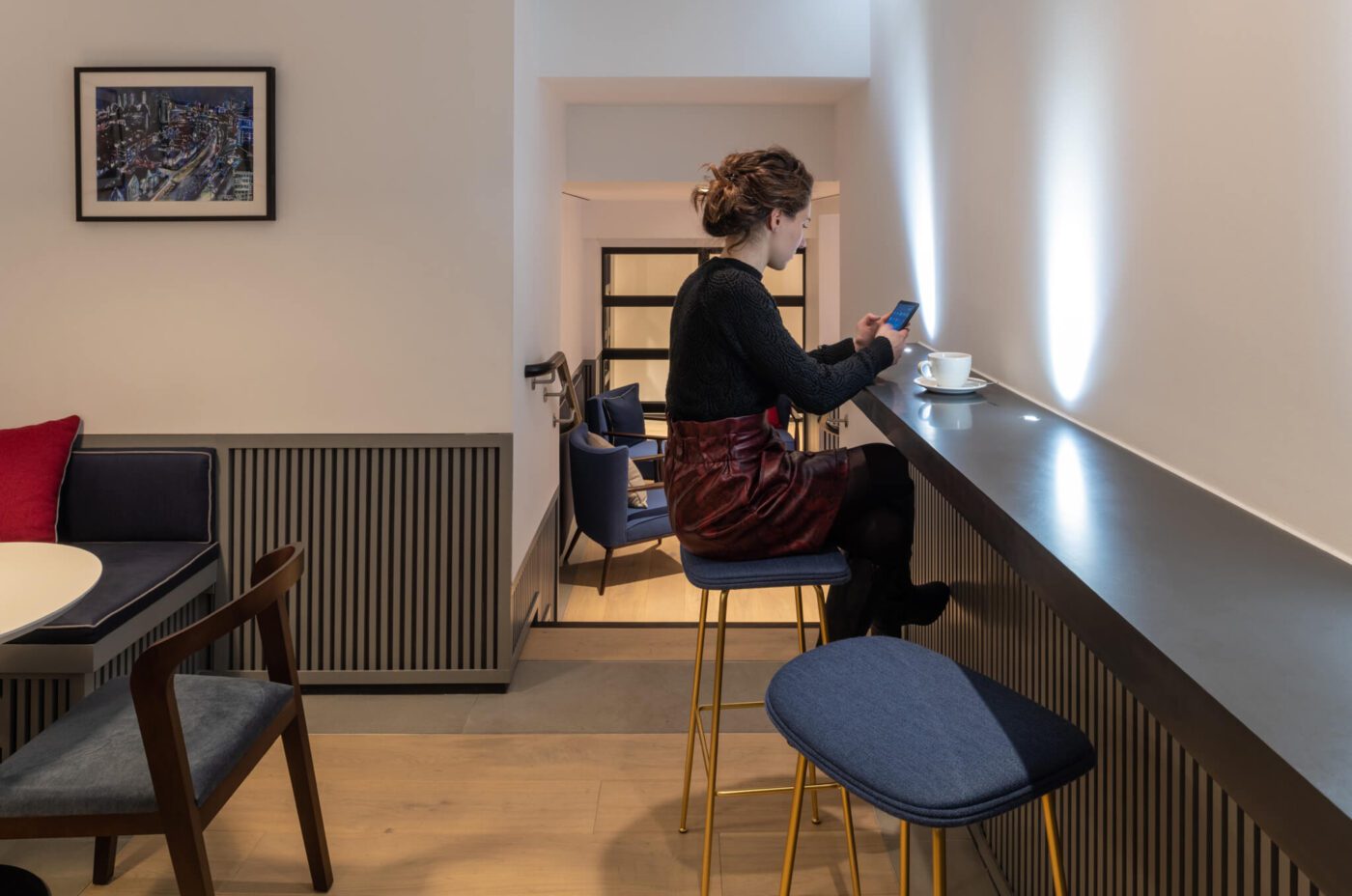 A person sits alone on a high stool at a counter in a modern, minimalist cafe. They are looking at their phone. There is a cup and saucer in front of them. Blue chairs and tables are visible in the background, along with a framed picture on the wall—a scene reminiscent of The Resident Covent Garden.