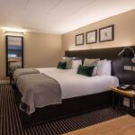 A modern hotel room at The Resident Liverpool features two neatly made twin beds with grey blankets and green pillows. Above the beds are three framed artworks. The room includes a large mirror, a wall-mounted TV, and a small wooden table. The floor is carpeted in a patterned design.