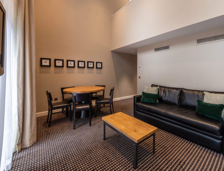 A modern hotel room at The Resident Liverpool featuring a black leather sofa with green cushions, a wooden coffee table, and a round dining table with four chairs. The walls are adorned with small framed pictures. The floor is carpeted, and there are tall curtains on the left side.