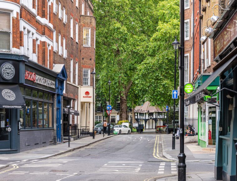 A quiet city street with various shops, including a pizza restaurant and a pub. The street is lined with brick buildings and has a few parked cars. A large green tree, reminiscent of the peaceful ambiance near The Resident Soho, adds a touch of nature to the urban setting.