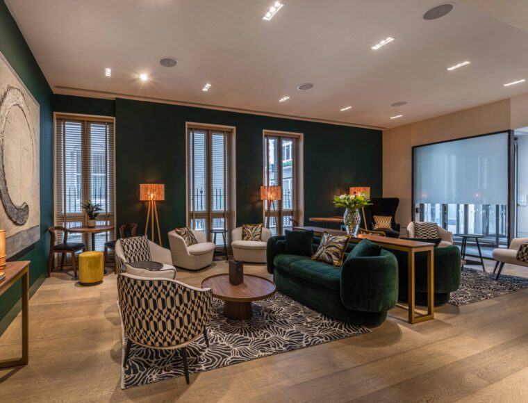 A modern, well-lit living room with green walls and wooden flooring, inspired by the elegance of The Resident Soho. It features a green velvet sofa, patterned chairs, a coffee table, and a large wall art piece. Tall windows with blinds line one wall. Several decorative lamps and a vase with flowers are also present.