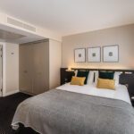 A modern bedroom at The Resident Soho features a large bed with white linens, mustard and green pillows, and a grey throw. Two bedside tables with lamps flank the bed. The room includes a large closet, a partially open door leading to a bathroom, and three framed artworks above the bed.