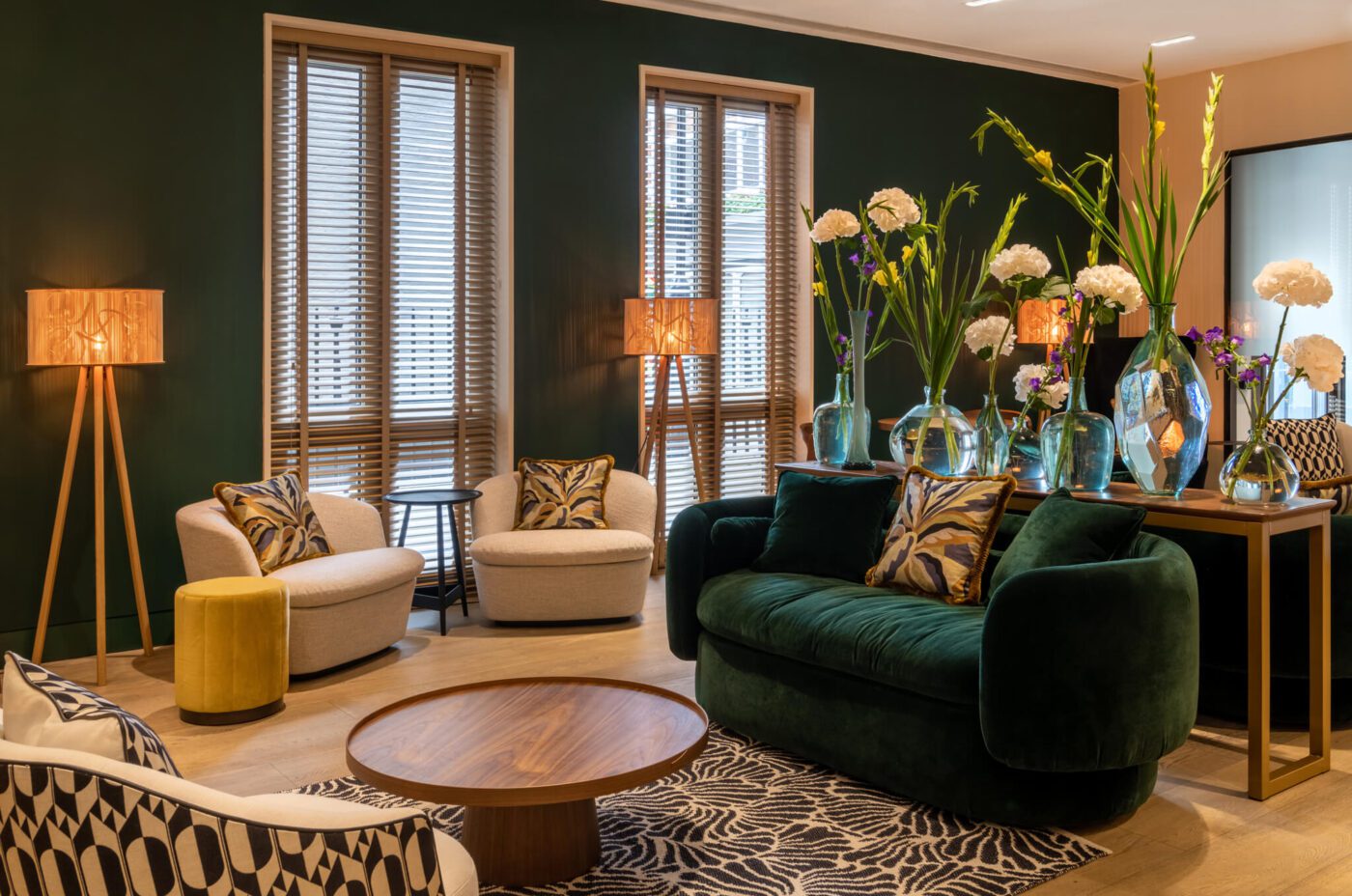 A modern living room with dark green walls and large windows at 4 star Soho hotel. The room features a green velvet sofa, beige chairs, a wooden coffee table, and a zebra-patterned rug. Vases of tall flowers sit on a console table while wooden floor lamps with decorative shades illuminate the space, reminiscent of The Resident Soho's chic decor.