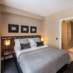 A neatly made bed with grey bedding and four pillows is situated in a modern hotel room at The Resident Victoria. This London hotel room has beige walls, two bedside lamps, a dark wooden headboard, and three framed pictures above the bed. A door on the right leads to a bathroom with a glass shower.