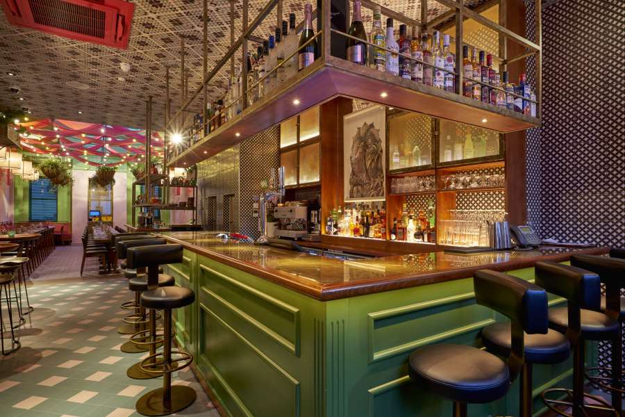 A chic bar interior with a green, L-shaped counter, black stools, and a metal overhead rack filled with bottles. The background features patterned walls, shelves with more bottles, and hanging plants. The space is well-lit with colourful ceiling decorations. The Cinnamon Bazar is a short walk from The Resident Covent Garden.