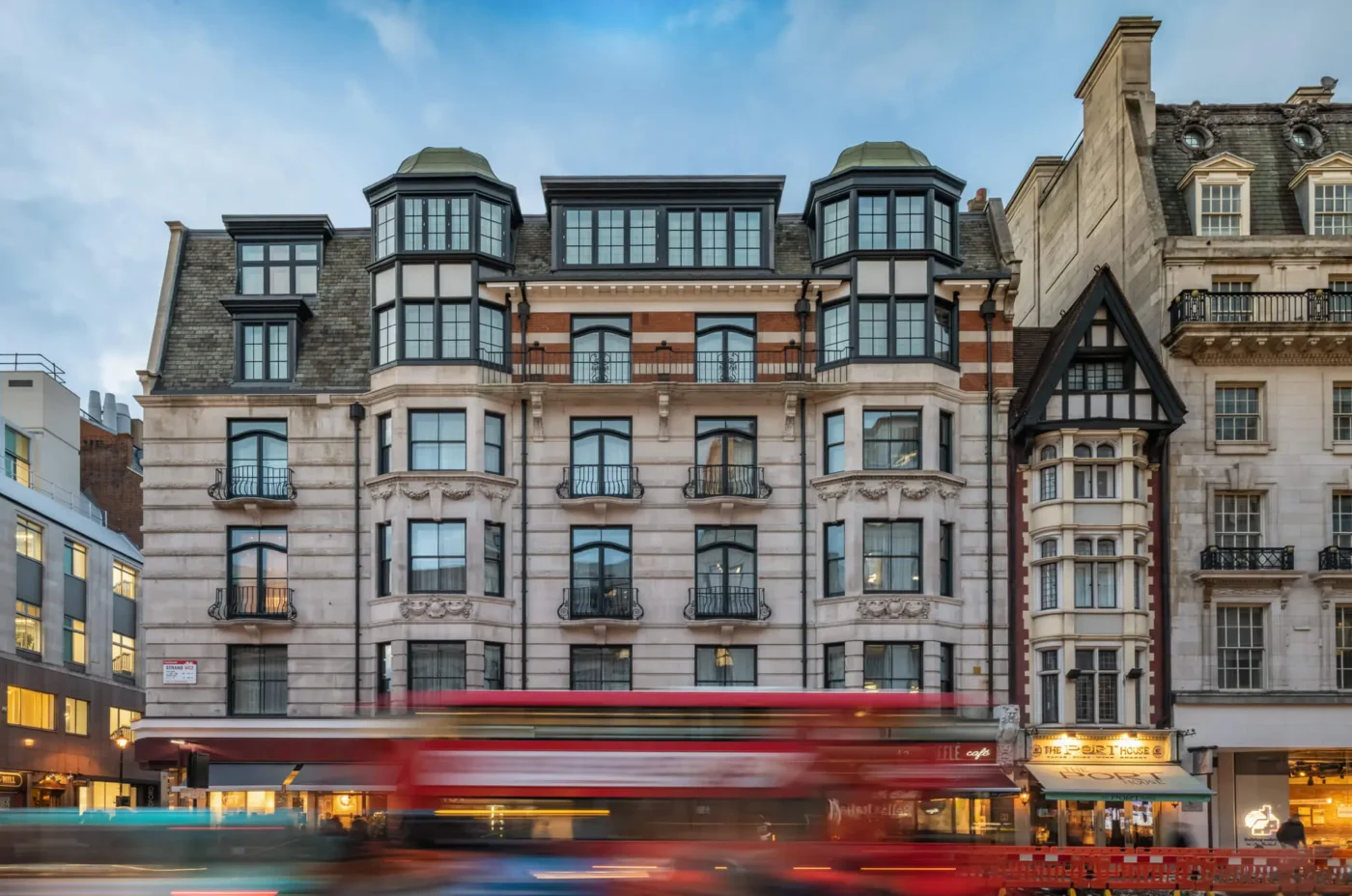 A wide shot of an ornate, multi-story building with bay windows and balconies, set on a busy city street. A red double-decker bus passes in front, creating a motion blur effect. Other historic buildings line the street.