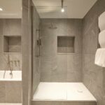 Modern bathroom with grey stone tiles featuring a glass shower enclosure. Inside the shower, there are two niche shelves. A separate bathtub with a handheld faucet is visible. Towels are neatly stacked on a rack outside the shower.