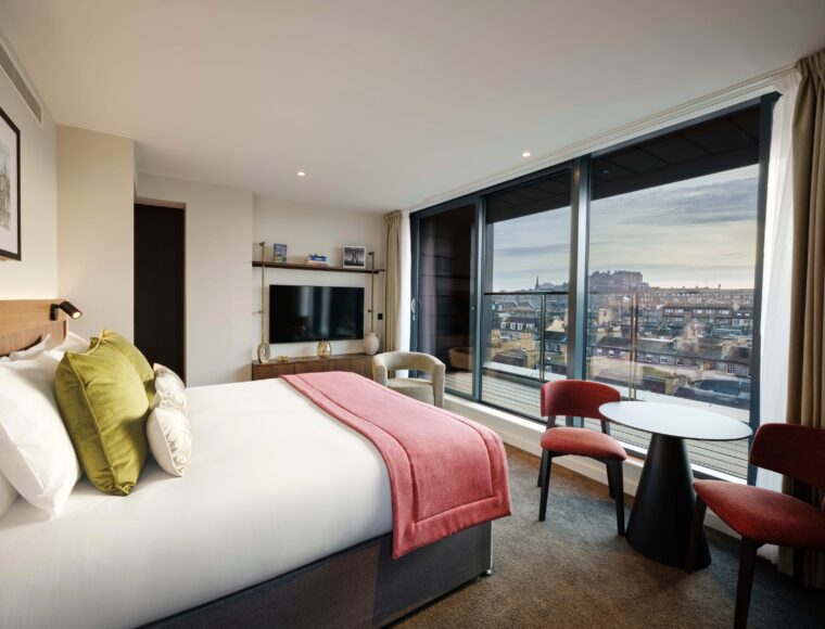 Hotel room with a large bed, red throw blanket, and decorative pillows. There's a TV on the wall, two red chairs, and a table by a large window showcasing a cityscape. Natural light streams in, highlighting the modern, cozy decor.