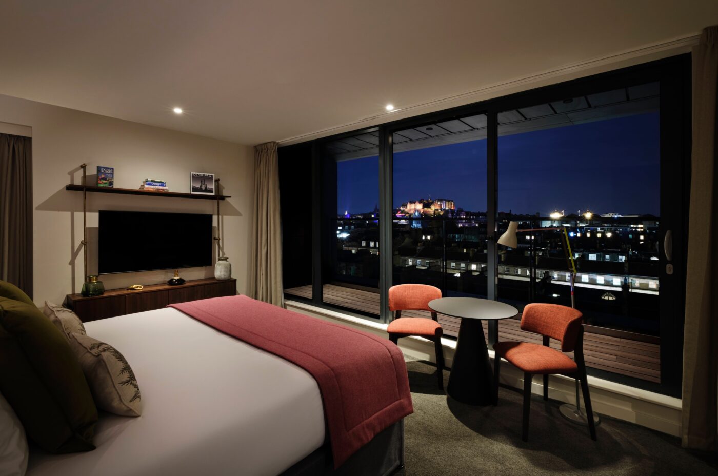 A modern hotel room with a large bed, pillows, and a pink throw. Two red chairs and a round table are near a large window offering a night view of a cityscape with illuminated buildings. A TV and shelves are mounted on the wall.