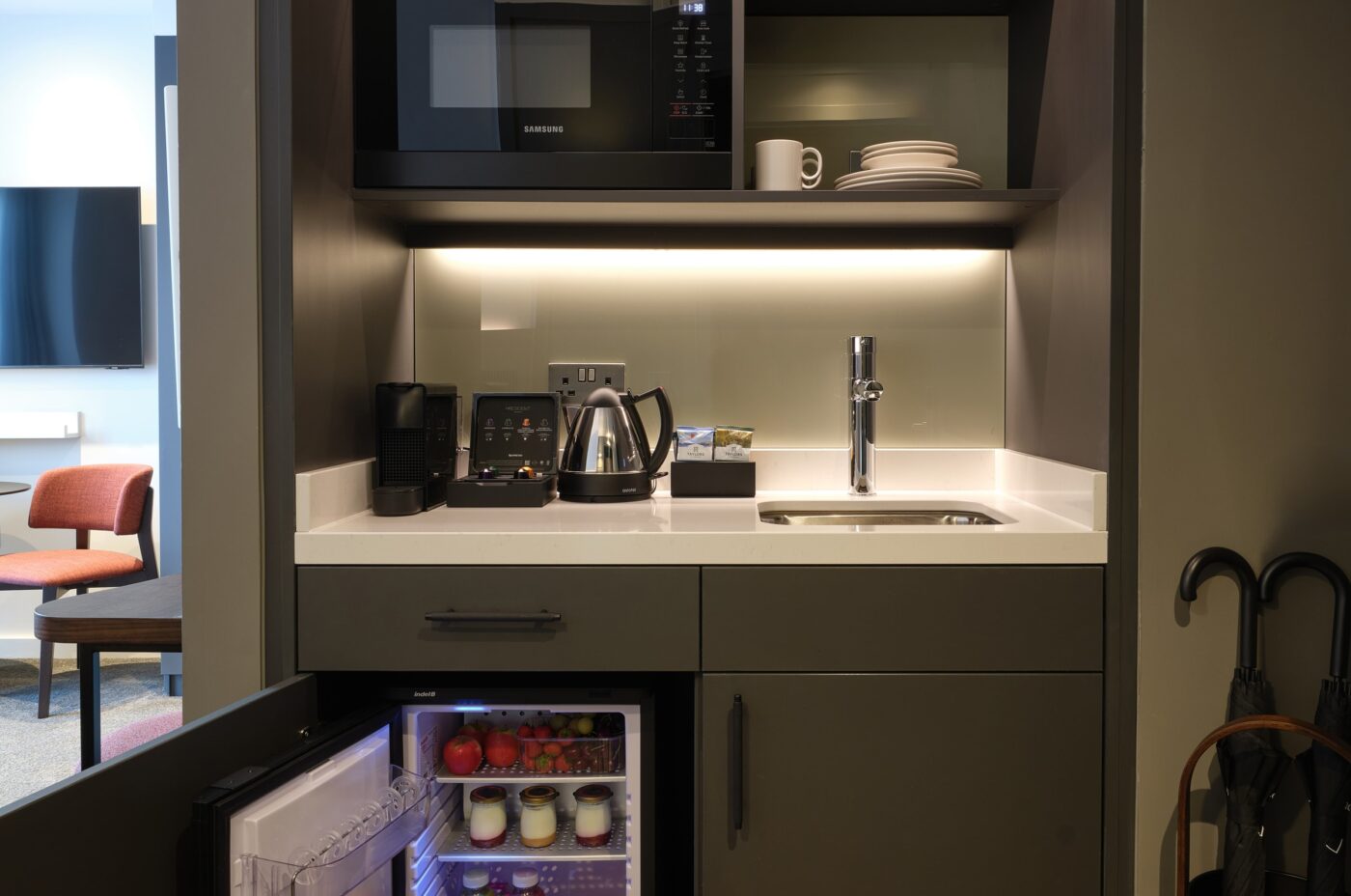 A compact kitchenette with a microwave, shelves with glassware, a sink, coffee maker, kettle, and a small fridge stocked with drinks and fruit. An umbrella stand is visible on the right.