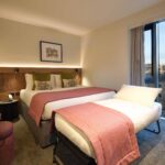 A hotel room with a large bed and a single pull-out bed, both with pink throws. A small desk and chair are in the corner. A window shows a cityscape outside. A framed picture hangs above the bed. The room is warmly lit.