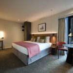 A modern hotel room with a large bed adorned with a pink throw and multiple pillows. There's a round mirror and a desk with stools on one side, and a small table with chairs by the window. Warm lighting and a framed picture create a cozy atmosphere.