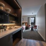 A modern hotel suite with a kitchenette on the left featuring a microwave, sink, and neatly arranged dishes. In the background, a cozy bedroom with a neatly made bed, armchairs, a table, and a large window with a city view.