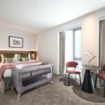 A bright hotel room with a large bed, a baby crib in front, two red chairs, and a round table with a lamp. The room has carpeted floors, two tall windows with curtains, and a framed picture above the bed.