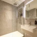 A modern bathroom with a glass-enclosed shower, featuring a rain showerhead. The walls and floor are covered in large gray tiles. A white sink with a sleek faucet is mounted on the wall, and a mirror above reflects the space.