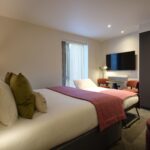 A modern hotel room with a cozy double bed featuring white sheets and pink accent throws. A flat-screen TV is mounted on the wall opposite the bed. There's a small seating area with a round table and chairs, and soft lighting creates a welcoming ambiance.