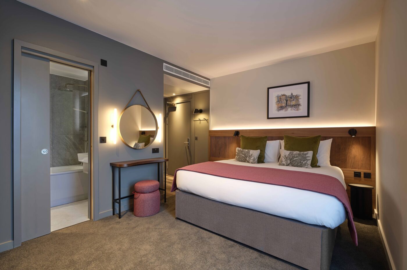 Modern hotel room with a large bed, pink throw, and green pillows. A round mirror hangs above a desk with a red cushioned stool. An open bathroom door reveals a bathtub. Soft lighting with carpeted floor and framed artwork above the bed.