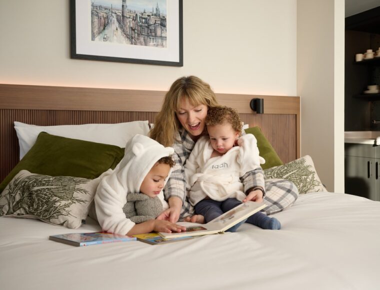 A woman and two small children sit on a bed. The woman is reading a book to the children, who are wearing cozy, hooded outfits. The bed is adorned with green pillows, and a framed picture hangs on the wall behind them.