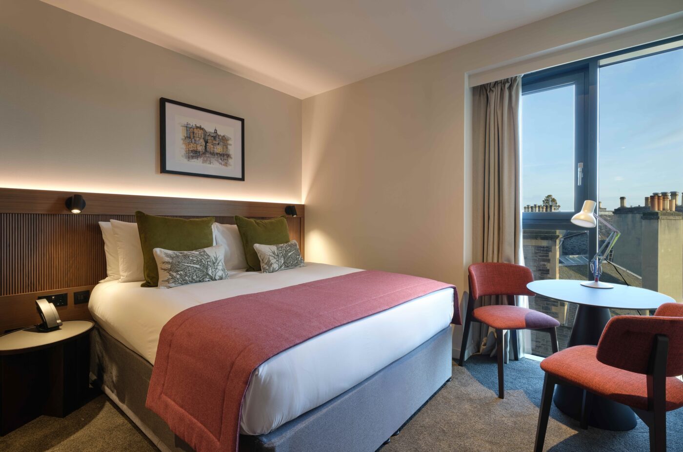 A modern hotel room with a king-sized bed featuring white linens, red and green cushions, and a red throw blanket. The room has two red chairs and a small round table by a large window with beige curtains, allowing natural light to enter.