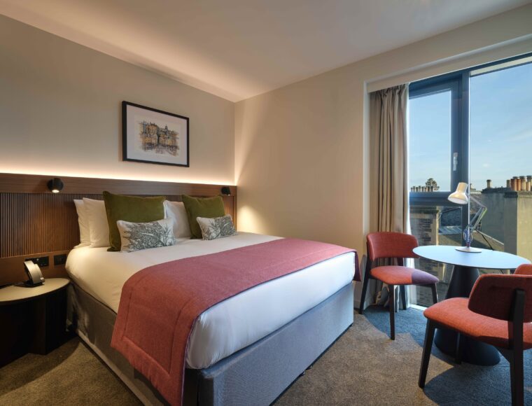 A modern hotel room with a king-sized bed featuring white linens, red and green cushions, and a red throw blanket. The room has two red chairs and a small round table by a large window with beige curtains, allowing natural light to enter.