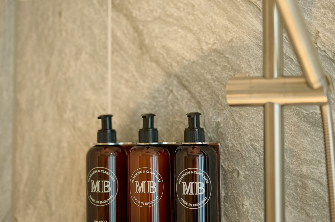 Three wall-mounted shower dispensers containing toiletries are installed against gray stone tiles. The dispensers have black bottles with pumps, labeled 