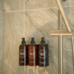Three sleek, dark-colored soap dispensers are mounted on a gray-tiled bathroom wall, featuring stylish branding. A silver showerhead on a vertical rod is visible nearby. The overall setting has a modern and minimalist design.