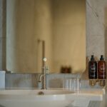 A modern bathroom sink with a rectangular mirror above it. To the right, there are two bottles of soap or lotion mounted on the wall. A tissue box and drinking glasses are placed on the sink. A metal grab bar is attached to the wall.