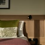 A cozy bedroom features a neatly made bed with white sheets, a green velvet pillow, and a decorative pillow with a botanical design. A maroon throw blanket covers part of the bed. A small spotlight and a framed picture adorn the wall.