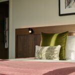 A cozy bedroom features a neatly made bed with green and patterned pillows, a pink blanket, and a wooden headboard. A framed picture hangs on the wall, and sunlight streams in from a nearby window, creating a warm atmosphere.