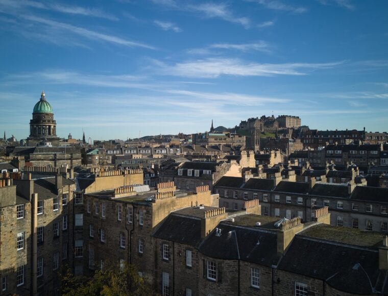 Aerial view of a historic cityscape with stone buildings under a blue sky. A prominent dome and an ancient castle are visible in the background, surrounded by rooftops.
