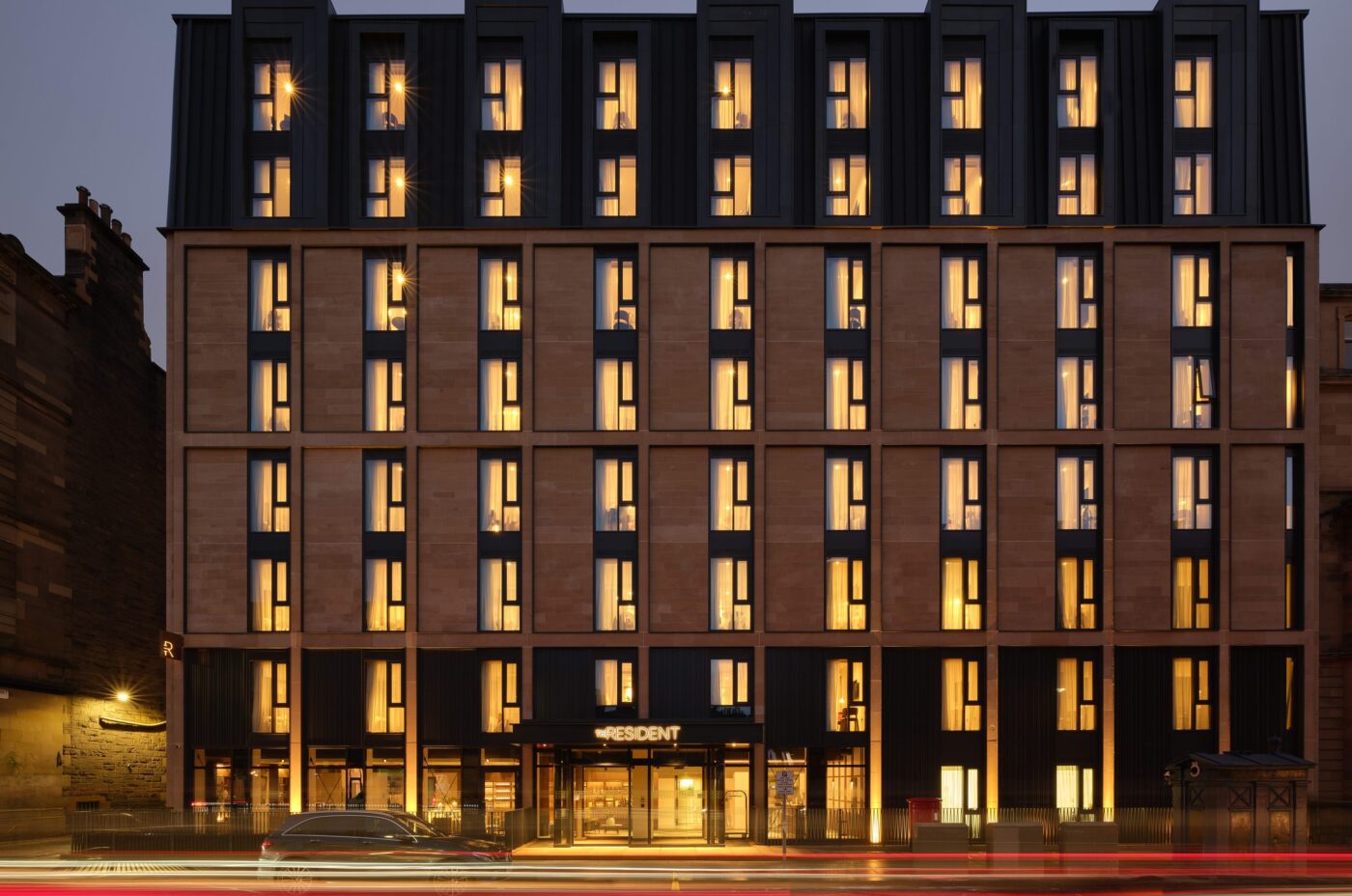 A modern hotel building with multiple stories is lit up in warm yellow tones against a dusky sky. The facade features vertical columns of windows, and a few cars drive past, creating light trails in the foreground.