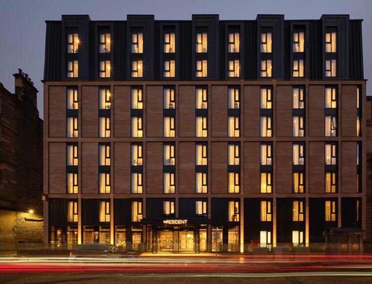 A modern hotel building with multiple stories is lit up in warm yellow tones against a dusky sky. The facade features vertical columns of windows, and a few cars drive past, creating light trails in the foreground.