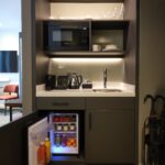 Compact kitchenette with a microwave, coffee maker, kettle, and sink set in dark cabinetry. An open mini-fridge below reveals drinks and fruits. Shelves hold glasses and plates. An umbrella is propped up nearby.