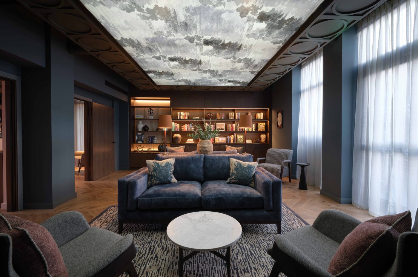 A modern living room with a dark, moody ambiance features a blue sofa, two gray armchairs, and a round marble coffee table on a patterned rug. The ceiling is artistically decorated. A wall lined with bookshelves and decorative items is in the background.