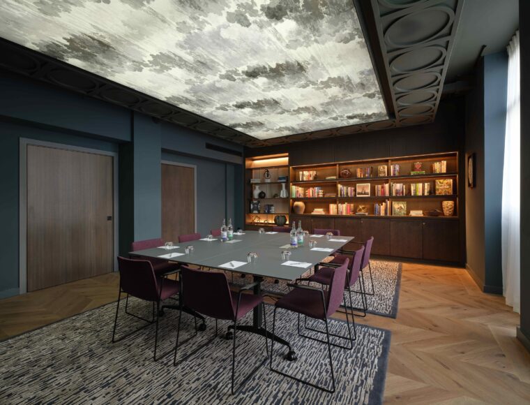 Modern conference room with a large rectangular table surrounded by maroon chairs. The ceiling features a cloudy design, and a wall has built-in shelves with books and decorations. Bottled water and glasses are placed on the table.