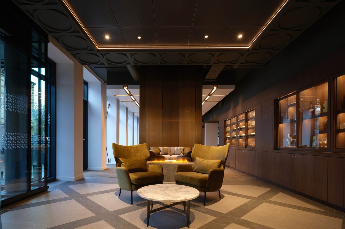 A modern, elegant lounge area with two velvet armchairs and a round marble table in front of a sleek fireplace. The room features large windows, wooden shelving with warm lighting, and a geometric-patterned floor.