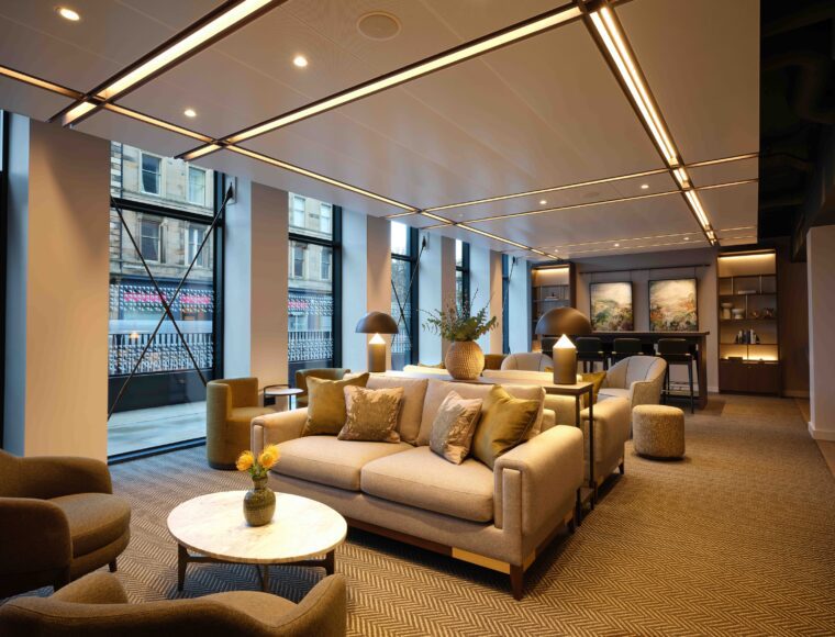 Modern lounge area with comfortable sofas and armchairs, decorated with cushions and plants. Large windows provide natural light, and a small round table with flowers is centered. A bar area with stools and artwork is visible in the background.