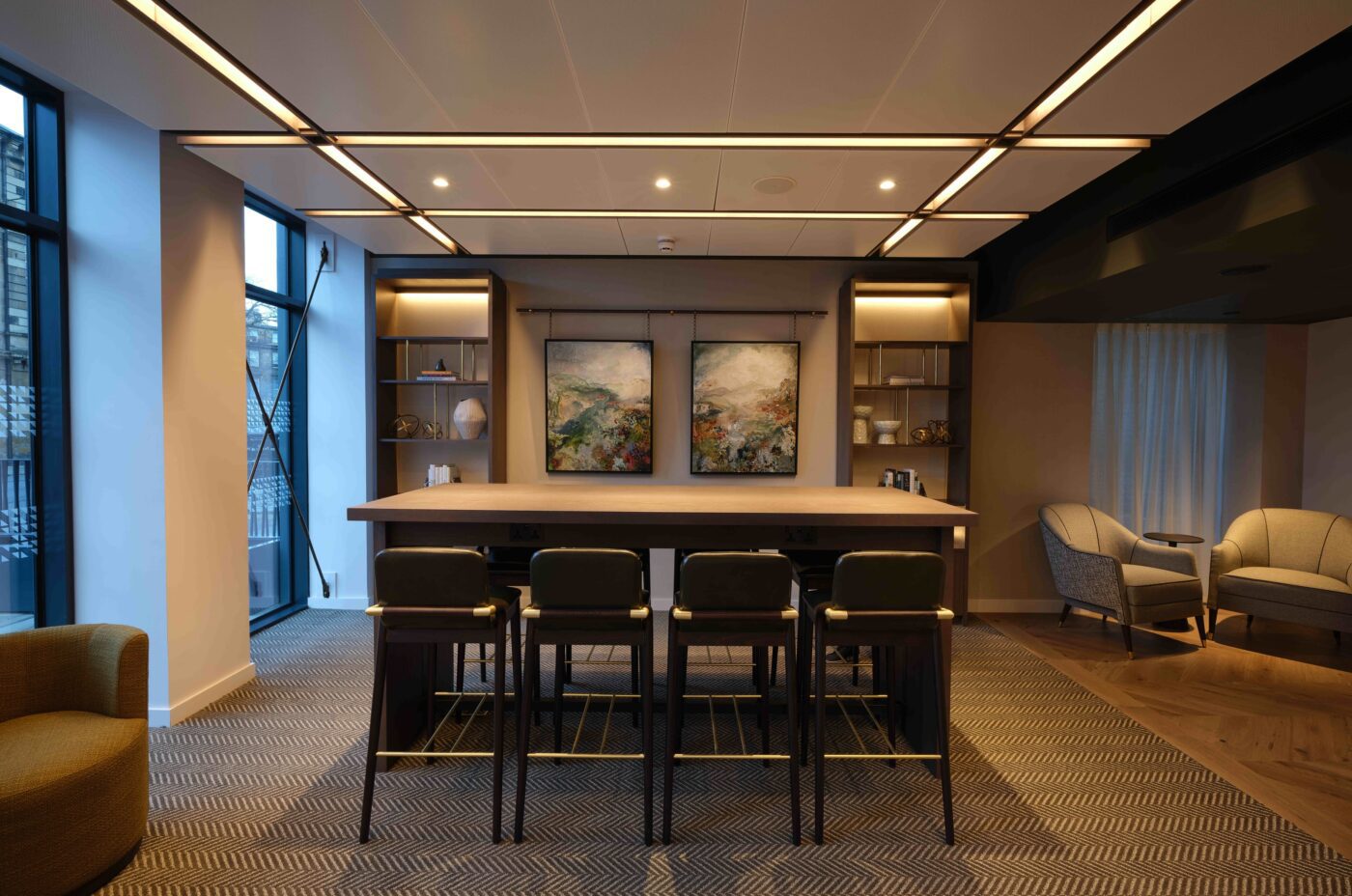 A modern office lounge with a long table surrounded by chairs, contemporary art on the wall, shelves with decorative items, and two armchairs in the corner. Large windows provide natural light, and recessed lighting adds a warm ambiance.