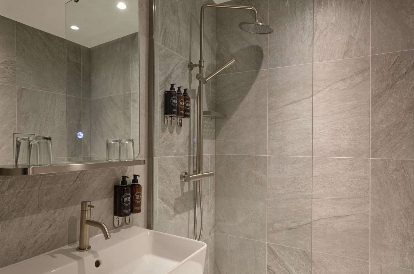 Modern bathroom featuring a walk-in shower with glass panels, a rainfall showerhead, and wall-mounted dispensers. A white sink with a metallic faucet, a large mirror, and gray stone tile walls complete the sleek design.