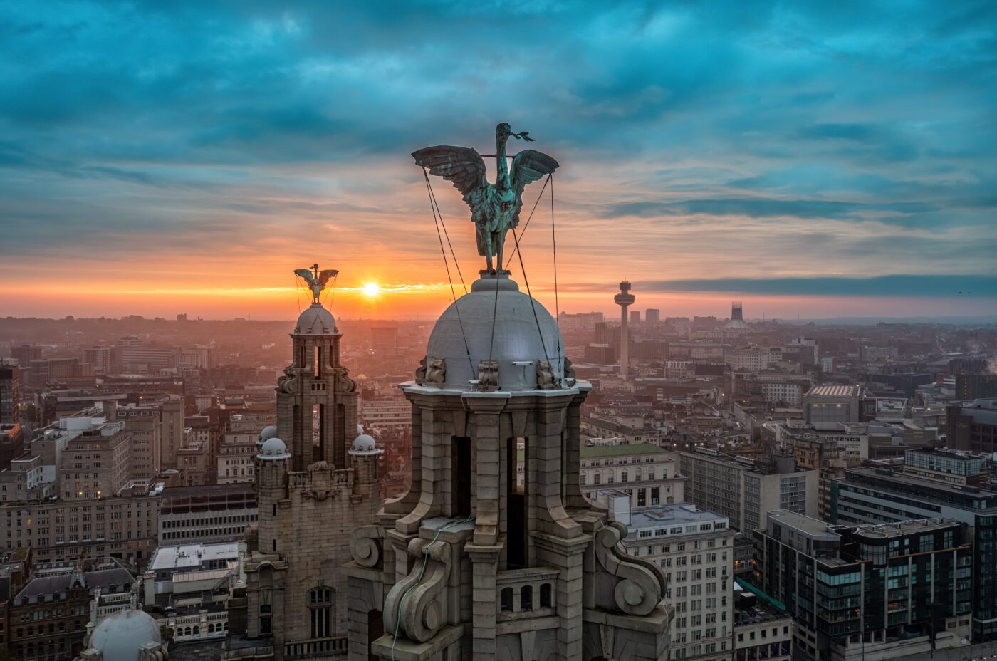 A cityscape view of Liverpool at sunset, featuring the Liver Building with its iconic Liver Bird statues on the domes, under a dramatic blue and orange sky.