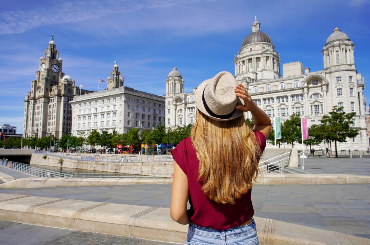 A woman with long blonde hair, wearing a hat and burgundy shirt, stands facing historic white buildings in Liverpool, UK, including the Royal Liver Building, under a blue sky.