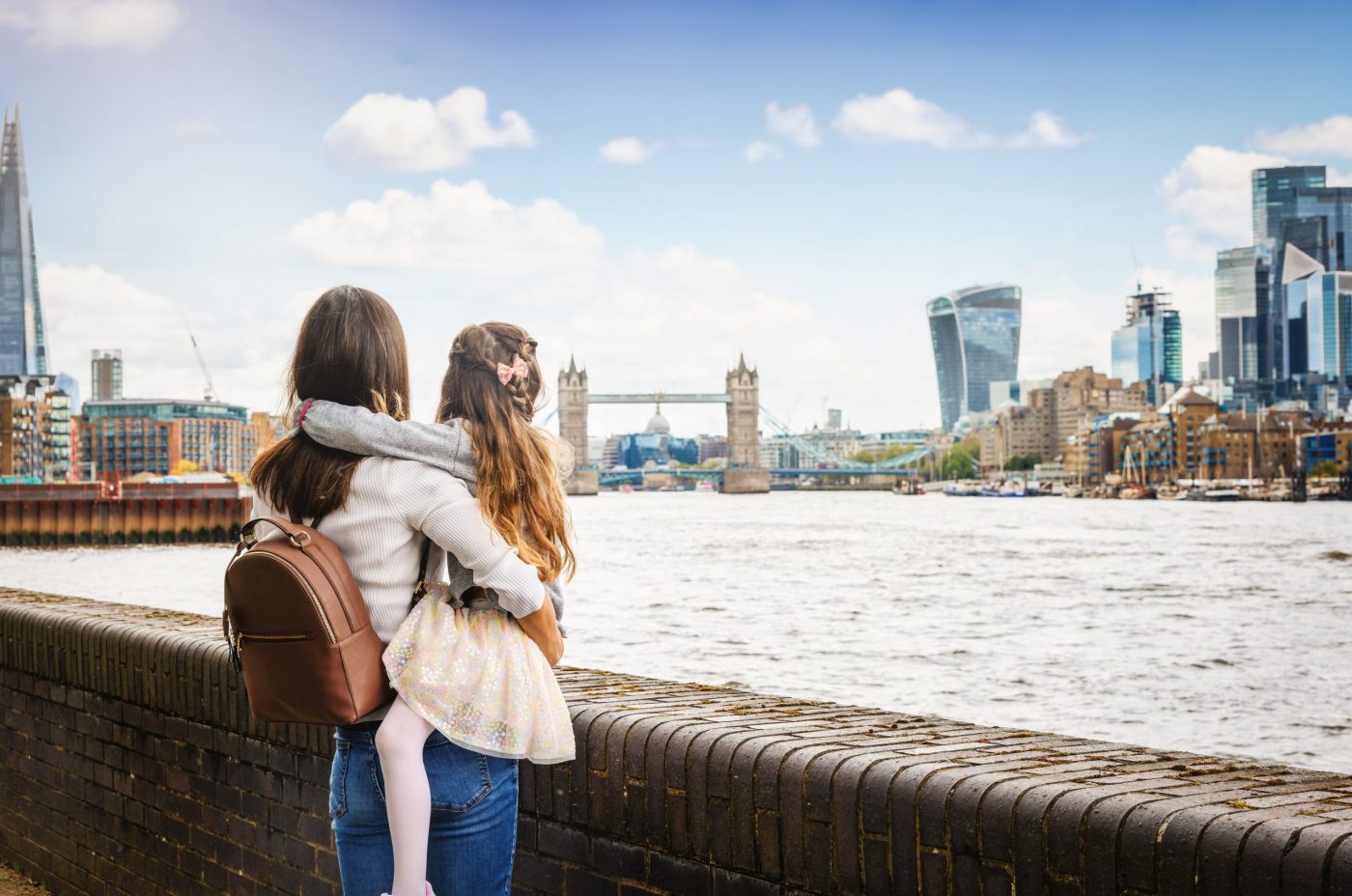 A woman with a brown backpack holds a young girl by the River Thames, looking toward the Tower Bridge and modern London skyline on a clear day.