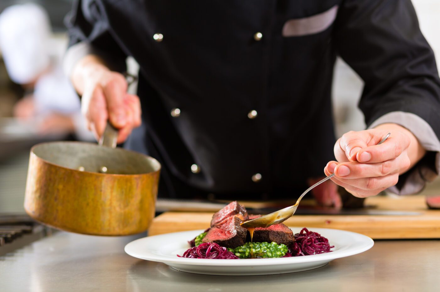 A chef in a black uniform carefully spoons sauce over a gourmet dish, featuring sliced meat, greens, and red cabbage as part of Liverpool Restaurant Week