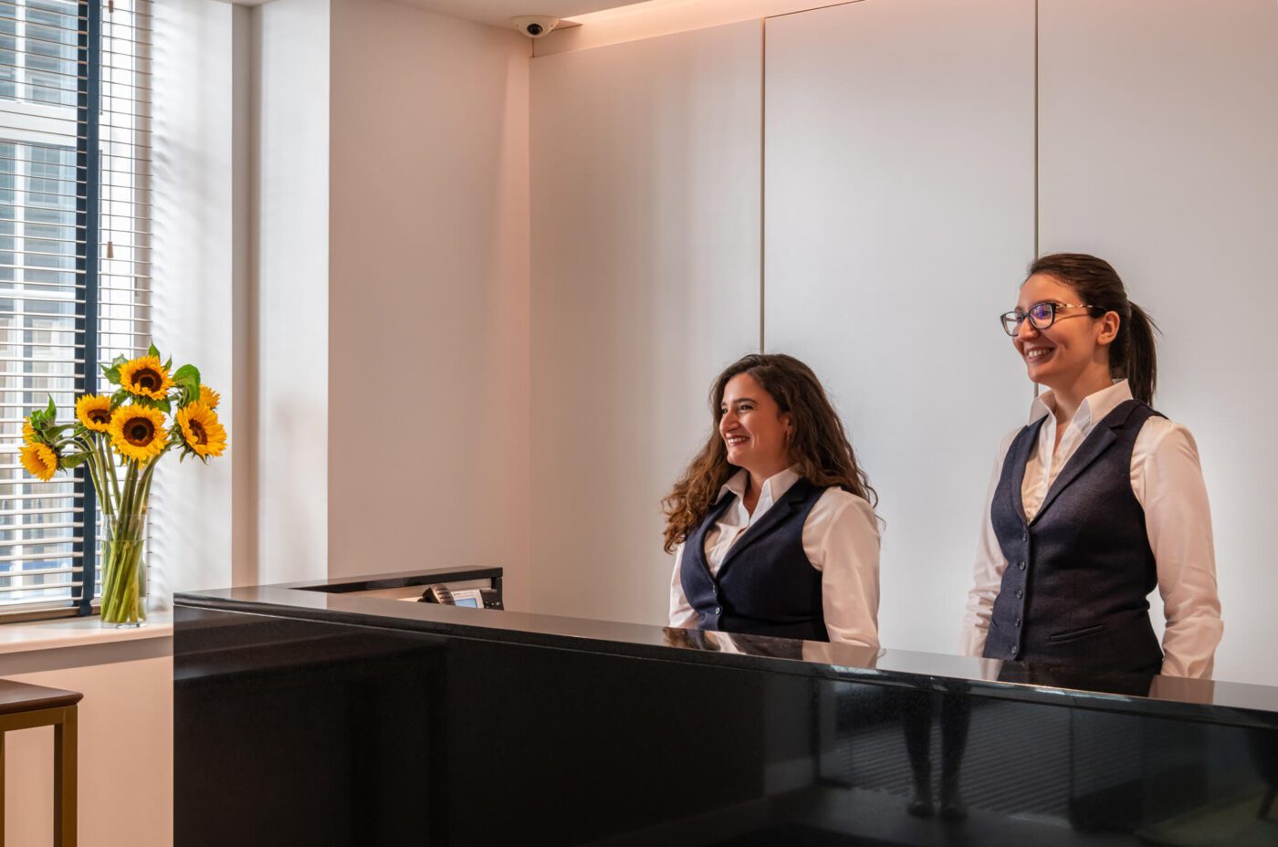 Two women in matching uniforms stand smiling behind a black reception desk in a modern office lobby. A vase of sunflowers sits on the windowsill to the left, and sunlight streams in through the window blinds.
