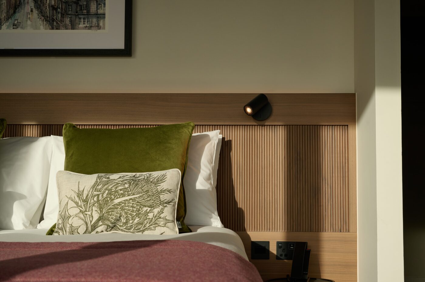 A neatly made bed with white sheets, a green pillow, and a decorative cushion featuring a botanical design sits against a wood-paneled headboard. A reading light is mounted above, and natural sunlight illuminates the scene.